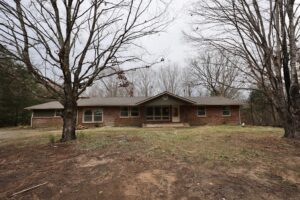 Single-story brick house with a front porch, surrounded by bare trees and a patchy lawn under an overcast sky.