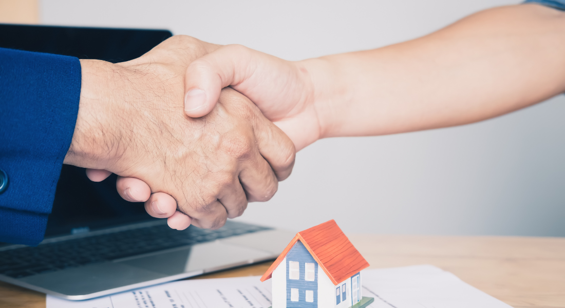 Two people shake hands over a desk with a small house model, documents, and a laptop, suggesting a real estate or business agreement.