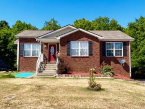 A small, single-story brick house with a red front door, concrete steps, and a patchy lawn, surrounded by trees under a clear blue sky.