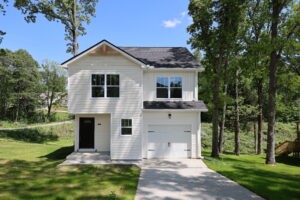 Two-story white house with black roof, attached garage, and black front door, surrounded by grass and trees on a sunny day.