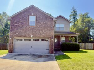 Two-story brick suburban house with a double garage, front porch, and well-kept lawn, set against a clear sky with trees in the background.
